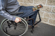 Outdoors by a light brick wall, a person in a wheelchair uses the Slim-Grip® Tablet Holder with Heavy-Duty Desk or Wheelchair Clamp Mount and 22 Arm to hold their tablet securely over their lap.