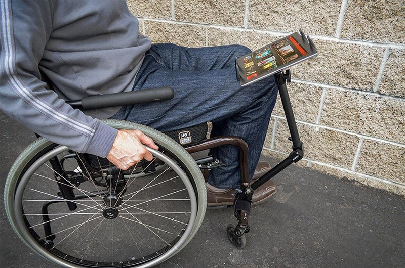 Outdoors by a light brick wall, a person in a wheelchair uses the Slim-Grip® Tablet Holder with Heavy-Duty Desk or Wheelchair Clamp Mount and 22 Arm to hold their tablet securely over their lap.