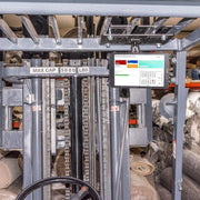 Close-up of a forklift with a 5000 lb capacity, equipped with the Slim-Grip® Tablet Holder with Overhead Forklift Guard Mount and 3.75 Shaft for warehouse management. Rolls of carpet are stored on shelves in the background.