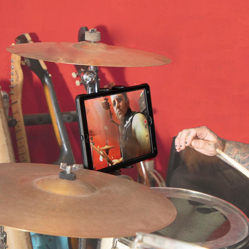 A person plays drums in a red-walled room with guitars behind. An iPad, held by a Slim-Grip® Tablet Mount with Clamp Base on the drum set, shows his focused face as he performs.