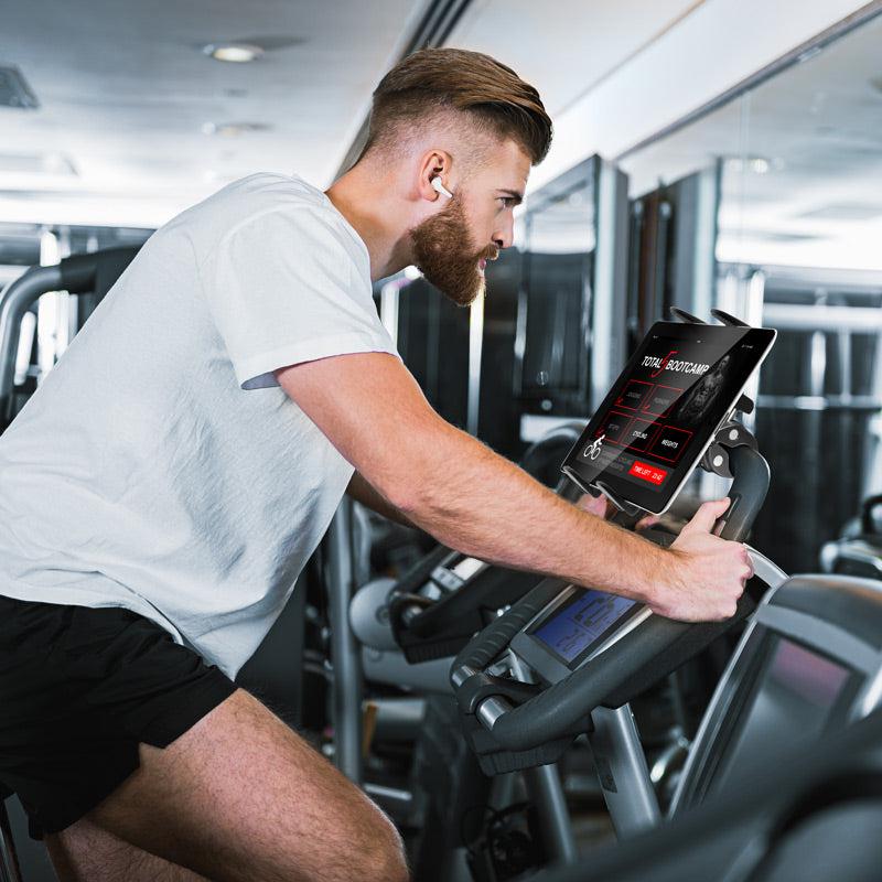 A bearded man in a white T-shirt and black shorts rides a stationary bike in the gym, viewing workout stats on a digital screen secured by the Slim-Grip® Tablet Mount with Clamp Base.