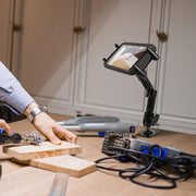 A person carves wood at a table, referencing a tablet held by the Slim-Grip® Ultra Heavy-Duty Multi-Angle Midsize Tablet Drilled-Base or Wall Mount, with a magnifying glass on a stand and various tools and cords scattered nearby.