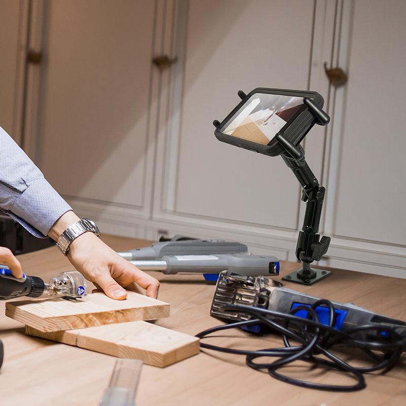 A person carves wood at a table, referencing a tablet held by the Slim-Grip® Ultra Heavy-Duty Multi-Angle Midsize Tablet Drilled-Base or Wall Mount, with a magnifying glass on a stand and various tools and cords scattered nearby.
