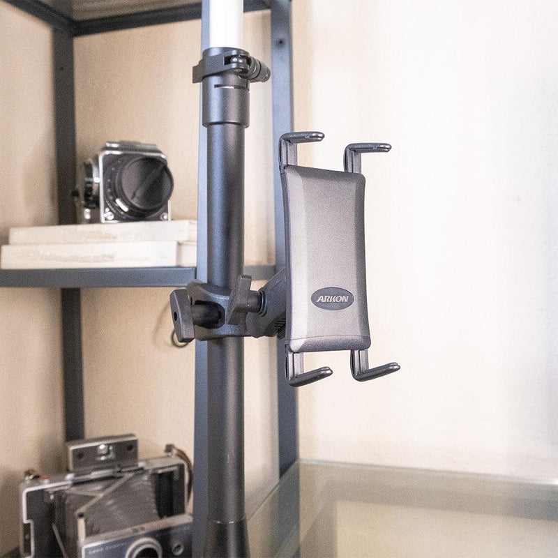 A Slim-Grip® Ultra Phone or Midsize Tablet Clamp Mount with Security Knob is attached to a vertical pole in front of a glass shelf, with vintage cameras and books visible on the shelves behind it.