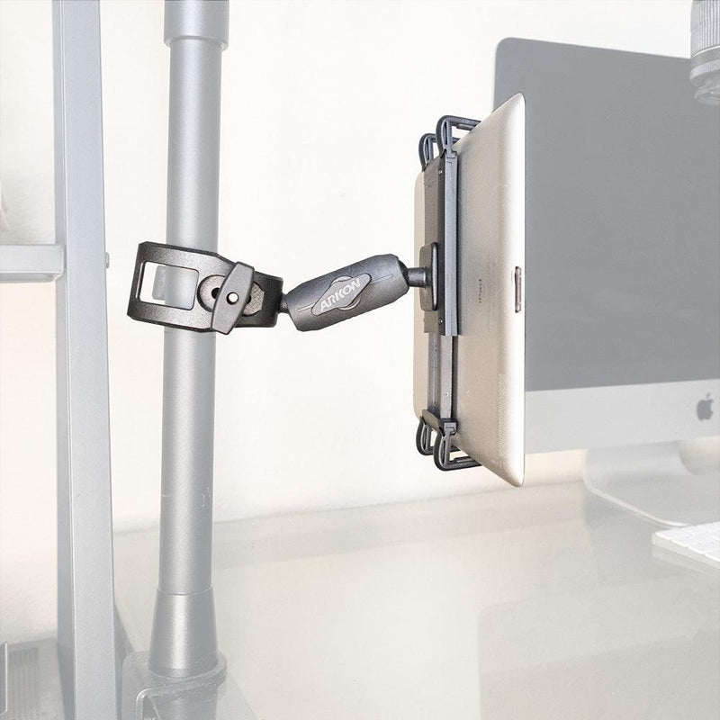 A Slim-Grip® Ultra Phone or Midsize Tablet Clamp Mount with Security Knob holds a tablet on a vertical metal pole beside a desk with a computer monitor in the background.