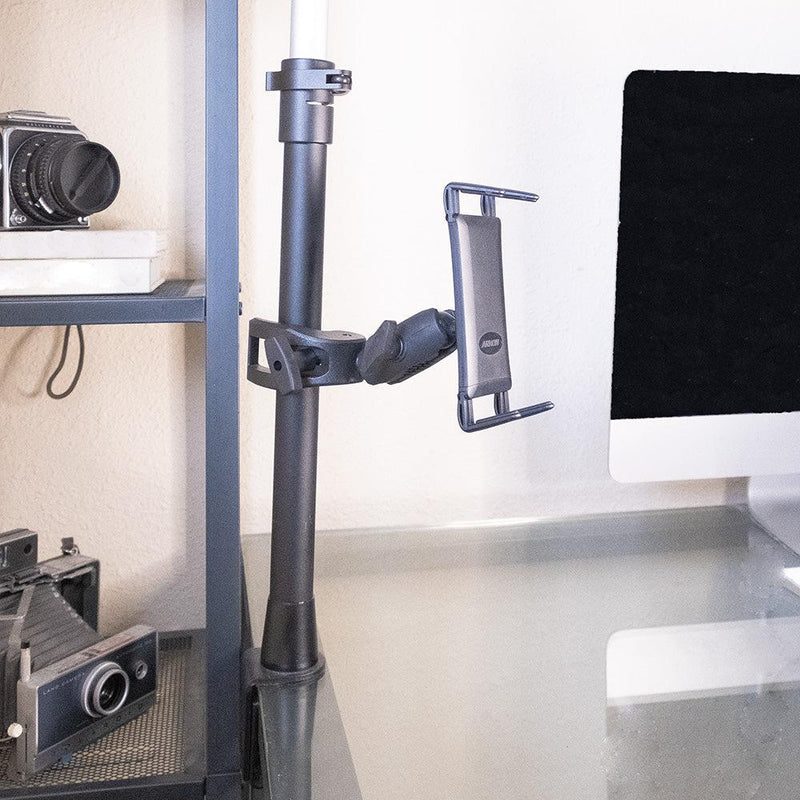A Slim-Grip® Ultra Phone or Midsize Tablet Clamp Mount with Security Knob is attached to a vertical pole beside a glass desk. Vintage cameras sit on a shelf nearby, and part of a computer monitor is visible on the right.