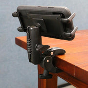 A Slim-Grip® Ultra Phone or Midsize Tablet Clamp Mount with Security Knob is attached to the edge of a wooden table, holding a smartphone in landscape orientation against a plain blue wall background.