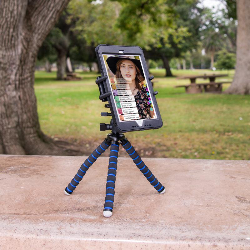 A tablet is attached to a blue and black Slim-Grip® Ultra Tripod Mount, set on a stone surface in a park. The screen shows a video call with a woman, and trees plus picnic tables are visible in the background.