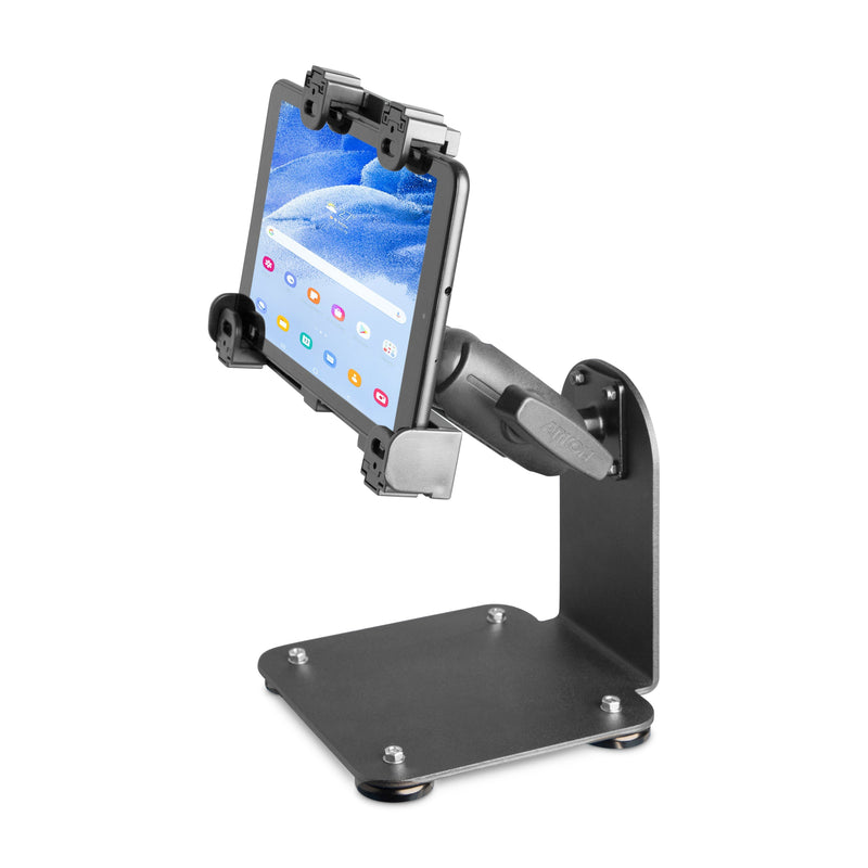 The SteadyMag™ Magnetic Mount System with LockVise™ Locking Tablet Holder securely holds a tablet displaying a beach scene. The mount features a weighted base and clamp mechanism, similar to a warehouse tablet stand.