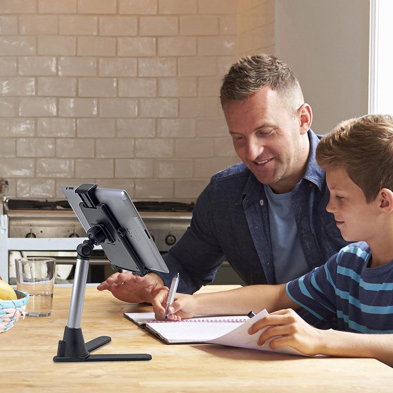 A man tutors a boy at a kitchen table as they use a Desk Stand Mount with Tablet Holder. The boy writes in a notebook while the man smiles. The kitchen has white tile walls and wooden surfaces.