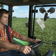 A man in a plaid shirt drives a green tractor through a vast field under a blue sky, using the Triple Robust Double Windshield Suction Mount - 17mm Ball Compatible fixed to the tractor’s window.