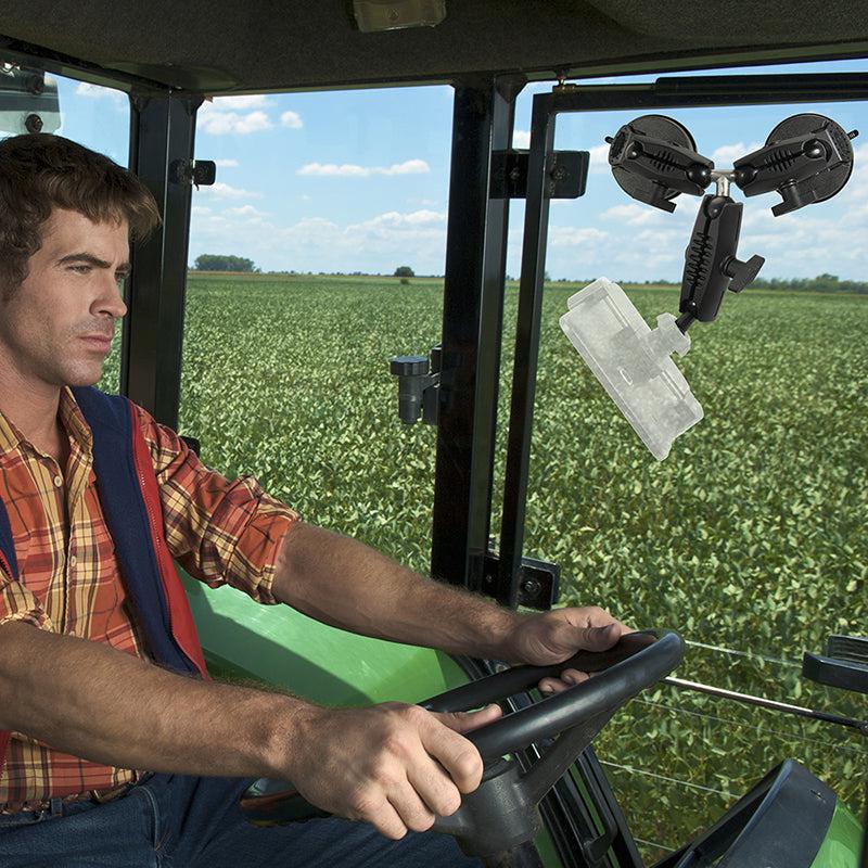 A man in a plaid shirt drives a green tractor through a vast field under a blue sky, using the Triple Robust Double Windshield Suction Mount - 17mm Ball Compatible fixed to the tractor’s window.