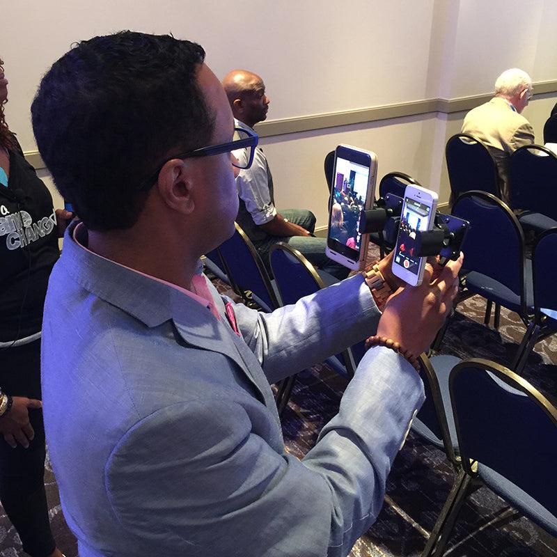 A man in a light blue suit films an event with the TW Broadcaster Pro - Side-by-Side RoadVise® Tripod Mount for Live Streaming on Facebook Live, using a handheld rig in a room with rows of blue chairs and several seated people.