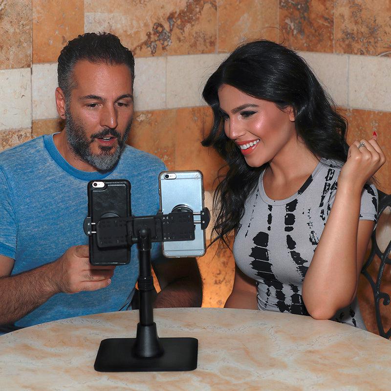 A man and woman sit at a table smiling, each looking at smartphones held by the TW Broadcaster Pro Stand – a Dual RoadVise® Phone Desk Stand ideal for live streaming on Instagram and Facebook Live, with a tiled wall in the background.