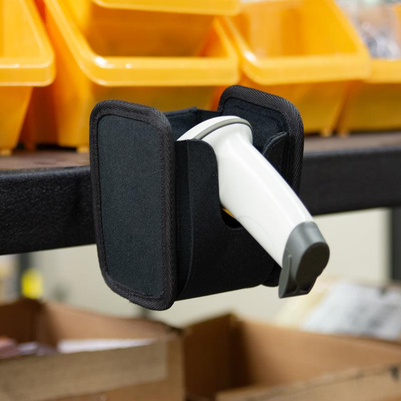 A white barcode scanner rests in a black Universal Barcode Scanner Holder on a shelf edge, with yellow storage bins and cardboard boxes behind it. The 4-hole AMPS compatible holder keeps the scanner securely in place.