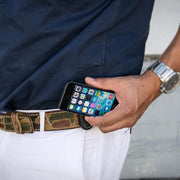 A person in a navy shirt and white pants wears a camo Universal Belt Clip Holder for Smartphones, with their phone showing apps. They also have on a silver wristwatch.